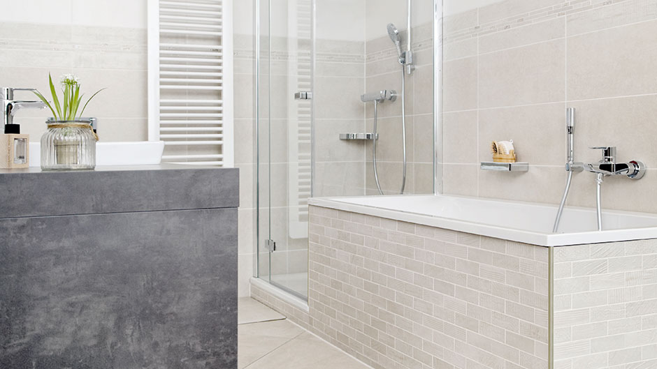 Das Badezimmer ist mit einem dunkelgrauen Waschtisch, beigefarbenen Wand- und Bodenfliesen sowie einer eingebauten Badewanne und einer barrierefreien Dusche mit Glastrennwänden ausgestattet.