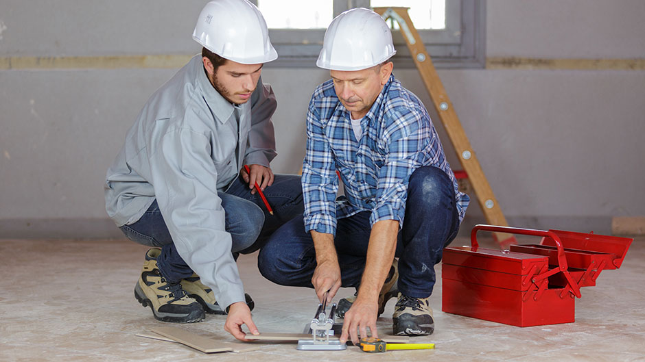 Zwei Handwerker sind damit beschäftigt, im Innenraum Fliesen zu verlegen.